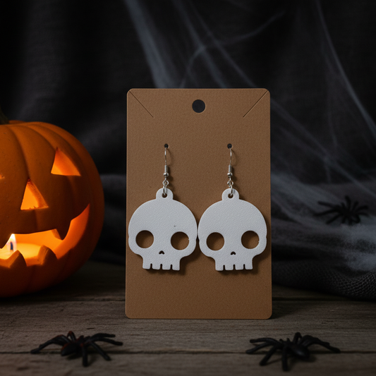 Skull-shaped earrings on a brown card with a Halloween-themed background featuring a jack-o'-lantern and spiders.
